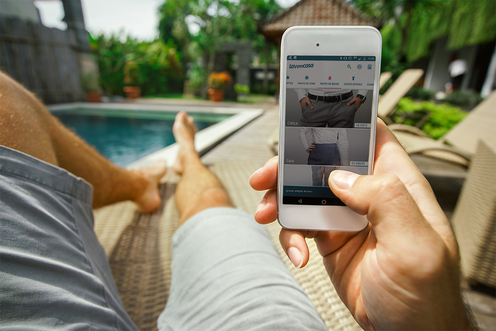 Uma pessoa relaxa em uma espreguiçadeira à beira da piscina, segurando um smartphone e navegando em uma loja de roupas online. A cena é ao ar livre, com vegetação e prédios ao fundo.