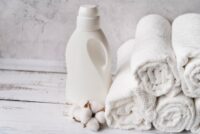 Uma garrafa de plástico branca fica ao lado de uma pilha de toalhas brancas cuidadosamente enroladas em uma superfície de madeira clara. Cápsulas de algodão estão espalhadas na frente, evocando limpeza e frescor. Perfeito para quem quer desencardir toalhas de banho, a cena contra a parede branca texturizada exala pureza.