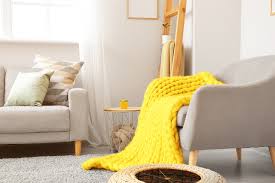 Uma sala de estar aconchegante com uma poltrona cinza coberta com um cobertor grosso de malha amarela, um sofá cinza claro com almofadas estampadas, uma pequena mesa lateral redonda e luz natural suave entrando por uma janela.