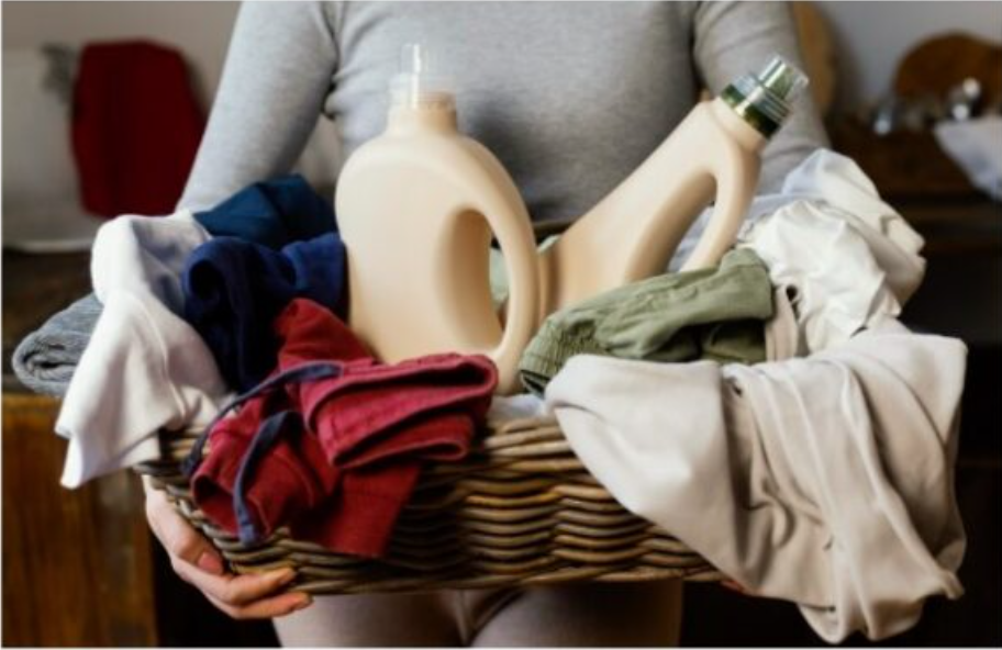 Uma pessoa em uma roupa cinza segura uma cesta de vime cheia de roupas dobradas, incluindo peças vibrantes vermelhas, brancas, verdes e azuis. Aninhadas entre as roupas e notavelmente roupas de lã estão duas grandes garrafas de detergente.