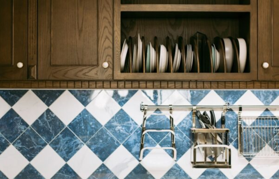 Uma cena de cozinha com armários de madeira com prateleiras abertas exibindo pratos empilhados. Abaixo, um backsplash com azulejos diamante azuis e brancos convida a pensamentos de como limpar azulejo de cozinha. Um suporte de metal segura utensílios e outras ferramentas essenciais de cozinha.