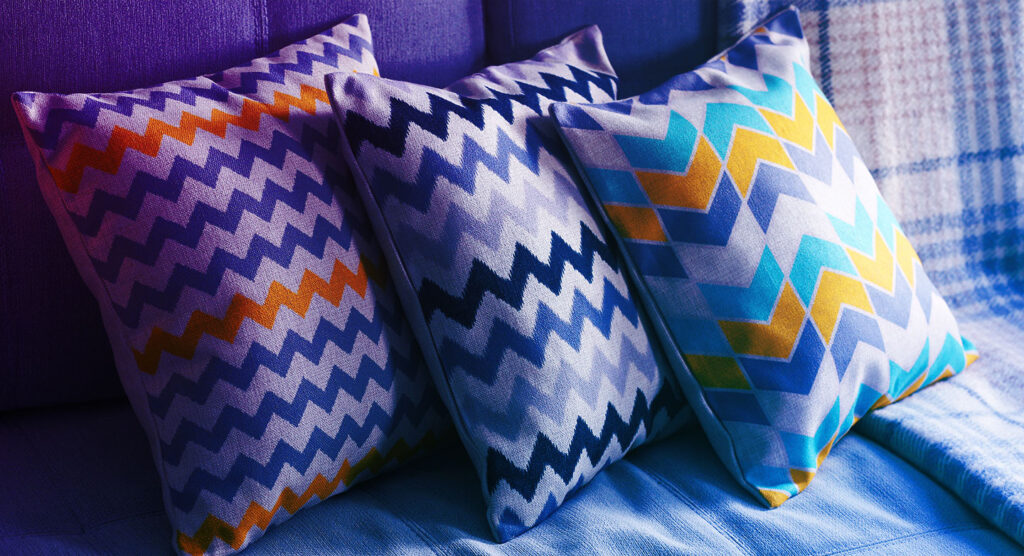 Três almofadas decorativas com padrões em ziguezague em tons de roxo, amarelo, preto e azul-petróleo estão dispostas em um sofá. O fundo apresenta um cobertor xadrez em tons frios semelhantes. A iluminação cria um ambiente suave e aconchegante.